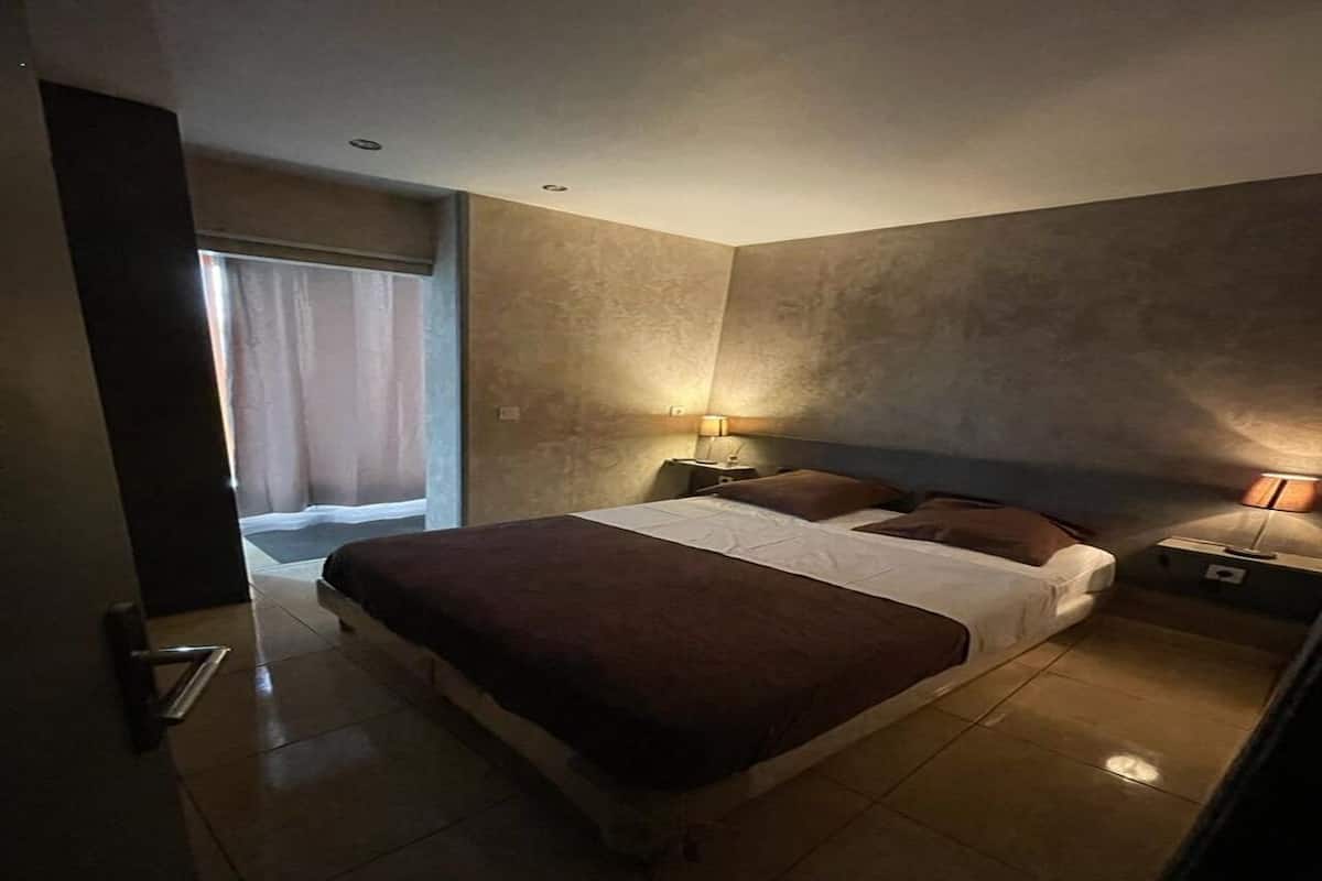 A bedroom with a large bed, two bedside tables with lamps, a door, and a view of a window with curtains.