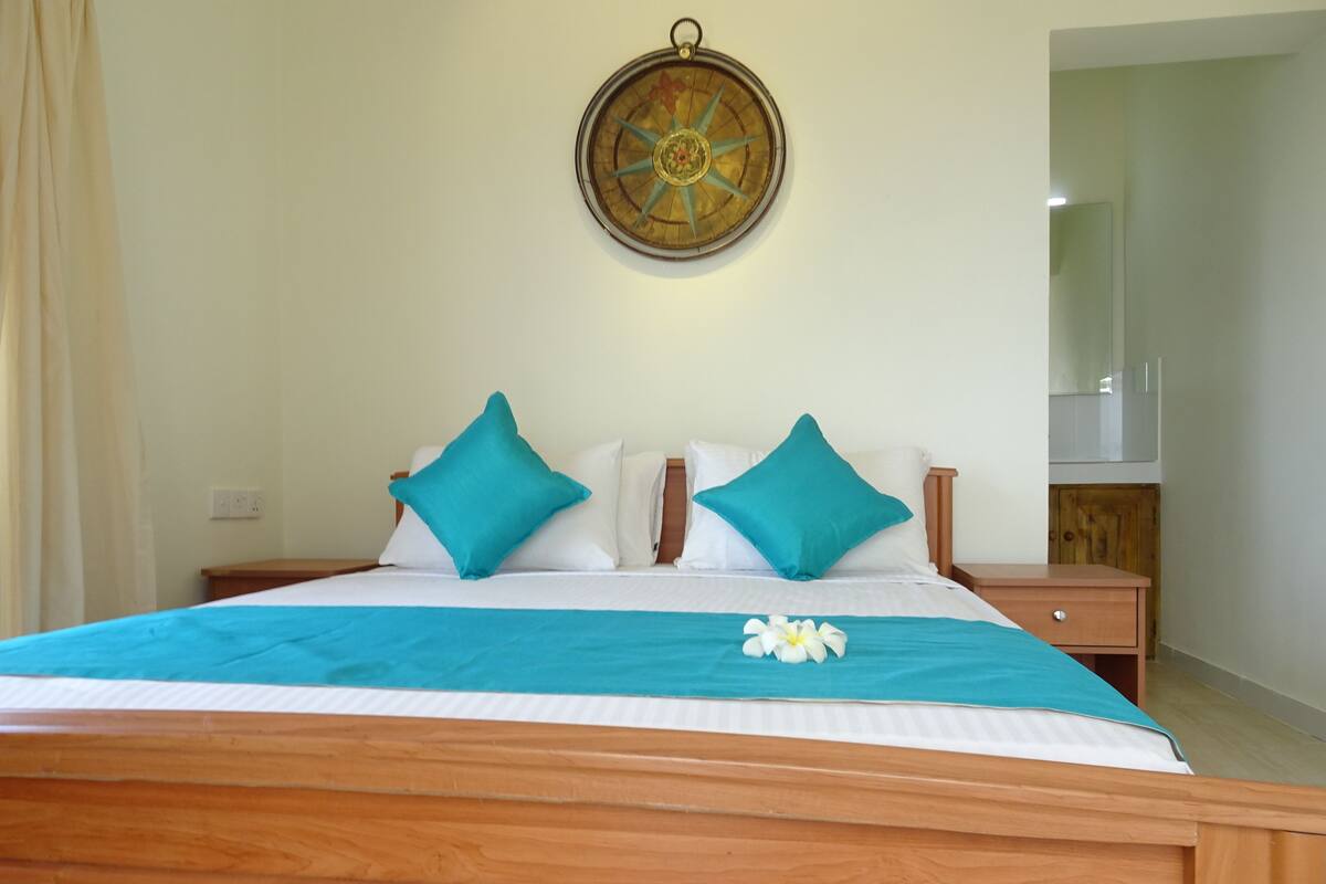 A neatly made bed with a teal bedspread and two teal pillows, a wooden headboard, and a wall-mounted compass.