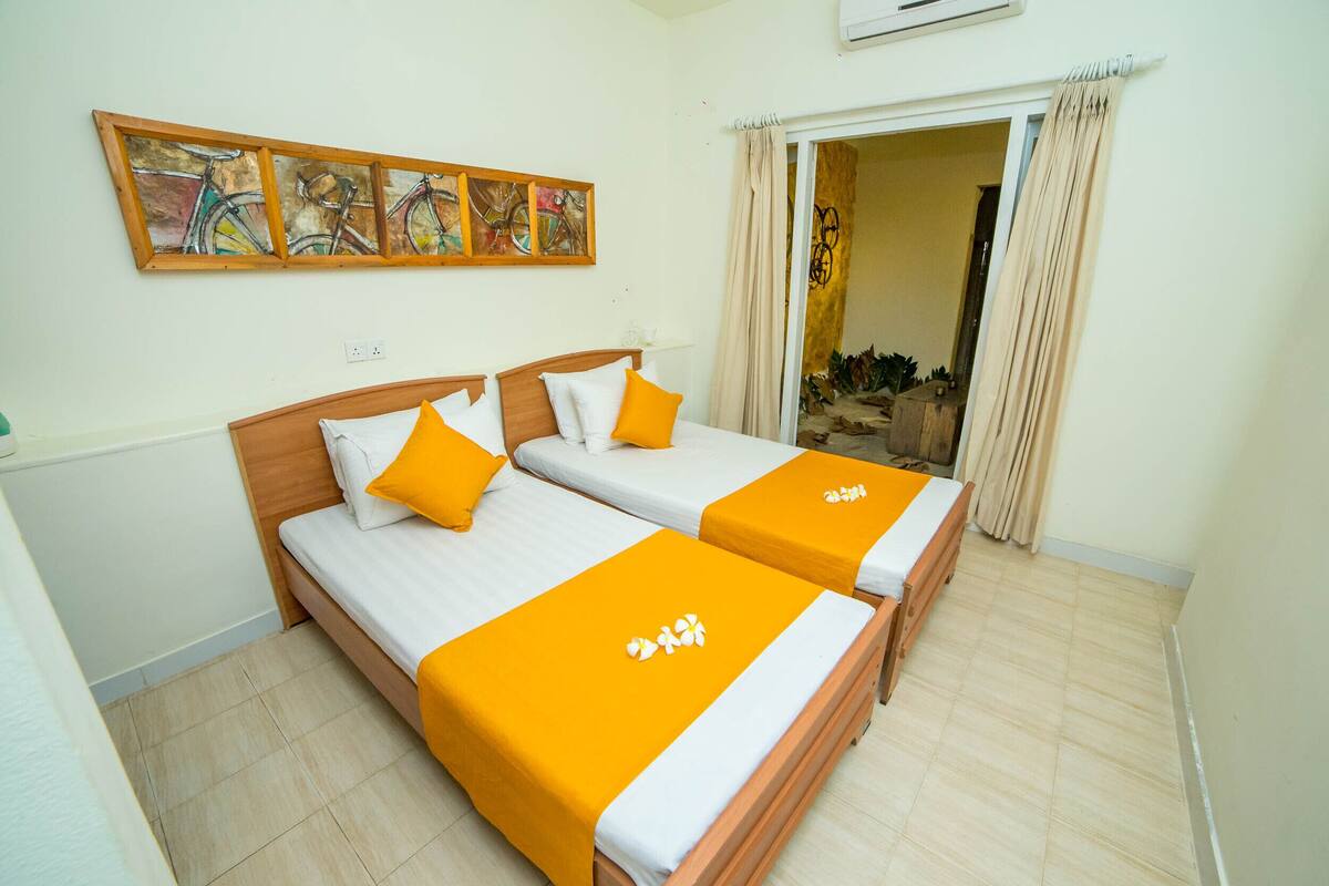 A hotel room with two beds, each with a wooden frame and white bedding, featuring orange bedspreads with a logo. There is a wall-mounted artwork above the beds and a door leading to a balcony with a view.