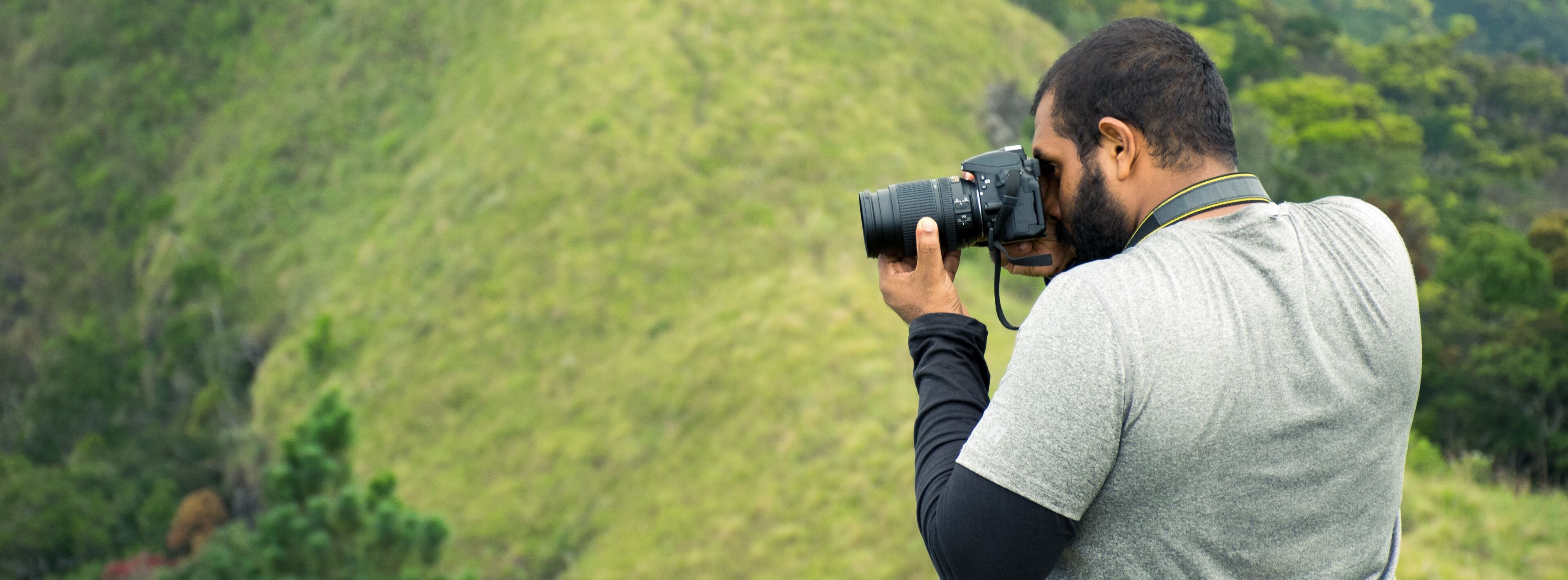 A person holding a camera, likely taking a photo, with a green, hilly landscape in the background.
