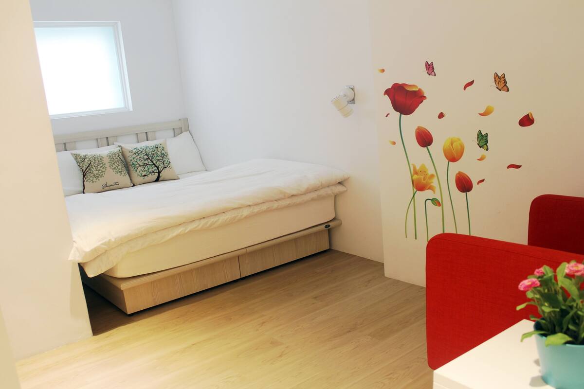 A bedroom with a bed, a window, a wall-mounted light, a red chair, and a small table with a plant.