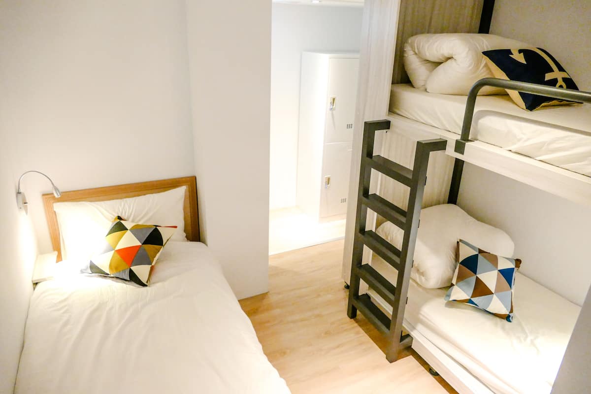 A bunk bed with a ladder, a single bed with a colorful pillow, and a bedside table with a lamp.