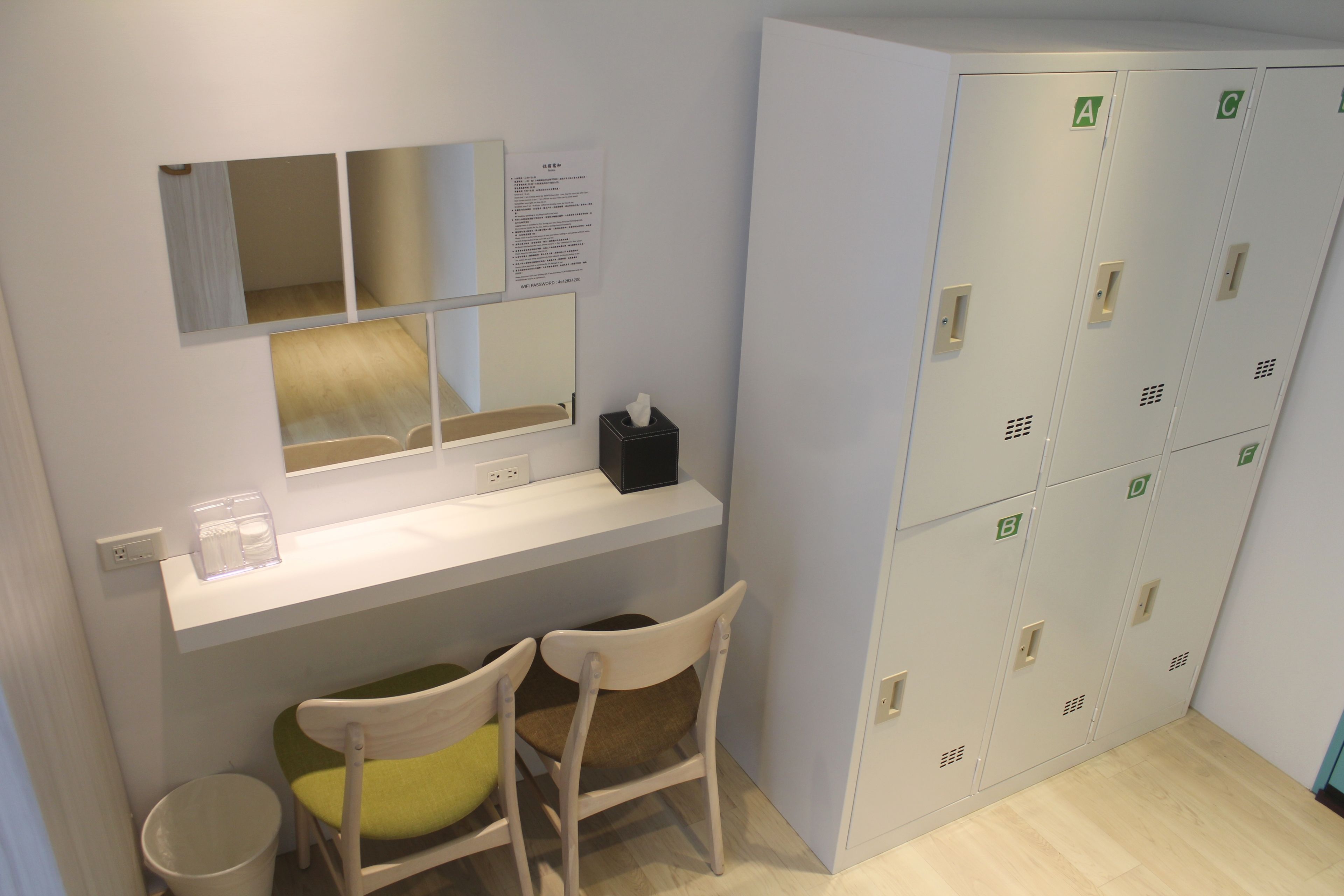 A small room with a desk, two chairs, a mirror, a bulletin board, and a set of lockers.