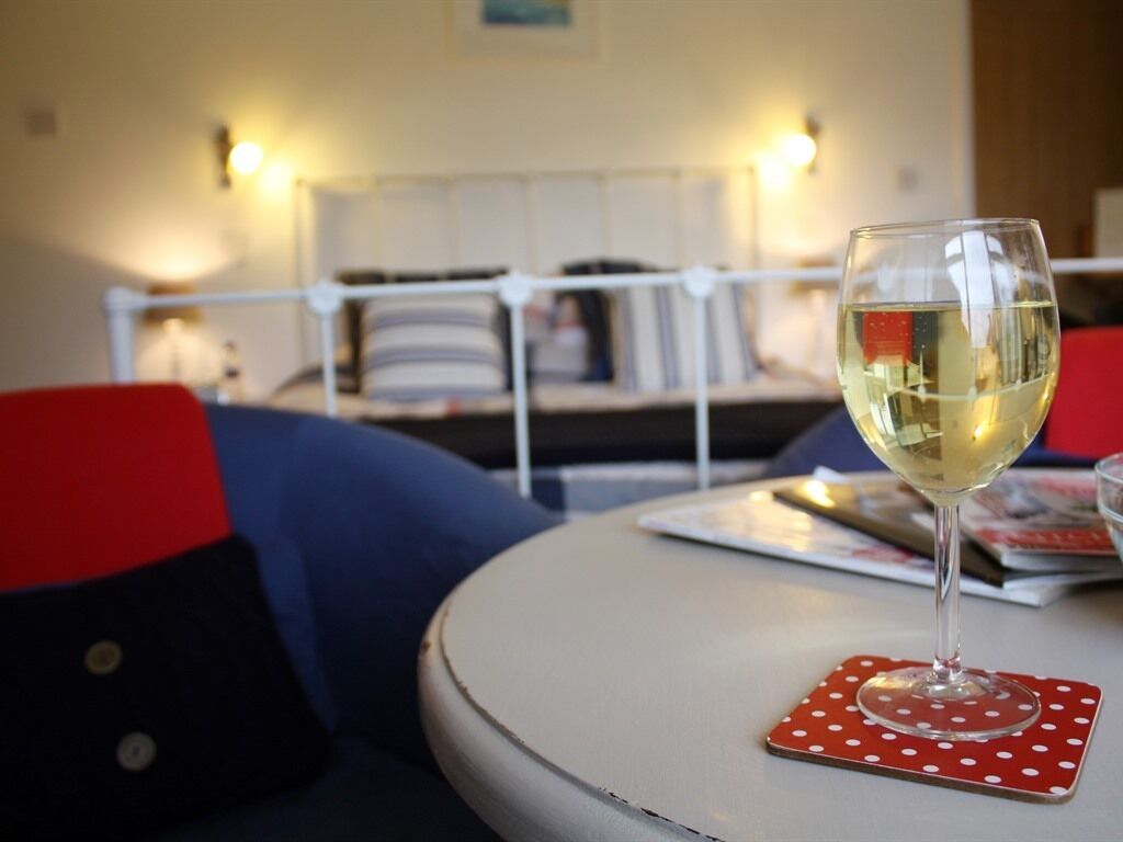 A round table with a glass of white wine on a red coaster with white polka dots. In the background, there is a bed with white headboard and pillows, and a blue chair.