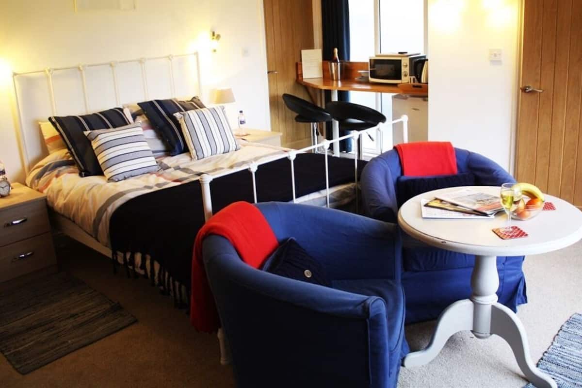 A compact bedroom with a bed, two chairs, a small table, and a kitchenette.