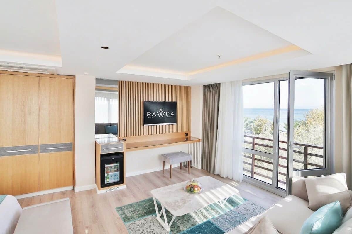 A modern hotel room with a kitchenette, a flat-screen TV, a sofa, a dining table, and a balcony with a view.