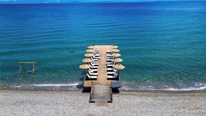 A wooden deck with lounge chairs and umbrellas, leading to a pebble beach. - Rawda Resort Hotel Altinoluk (Edremit)