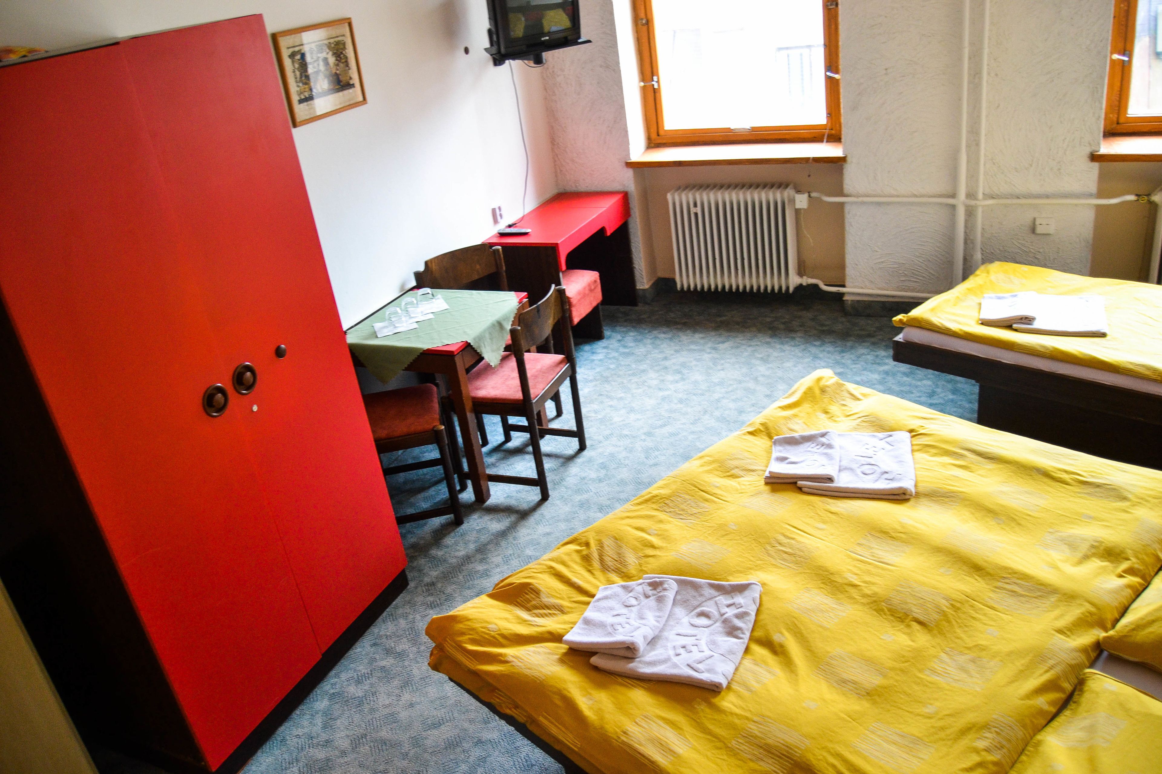 Una habitación con dos camas, un armario rojo, una mesita con mantel verde y una ventana con vistas.