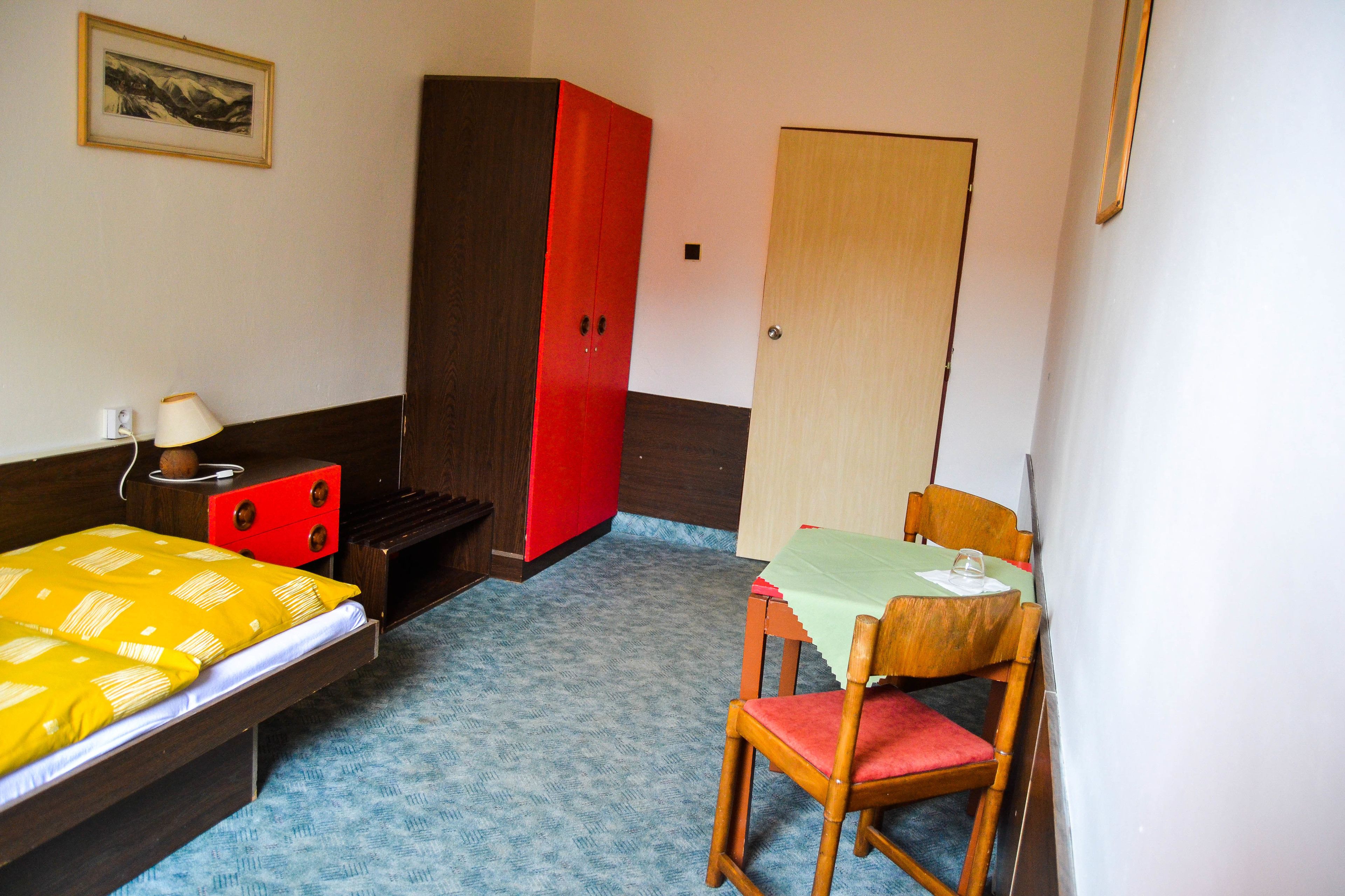 Un dormitorio con cama, un armario rojo, una silla de madera, una mesita con mantel verde y una puerta.