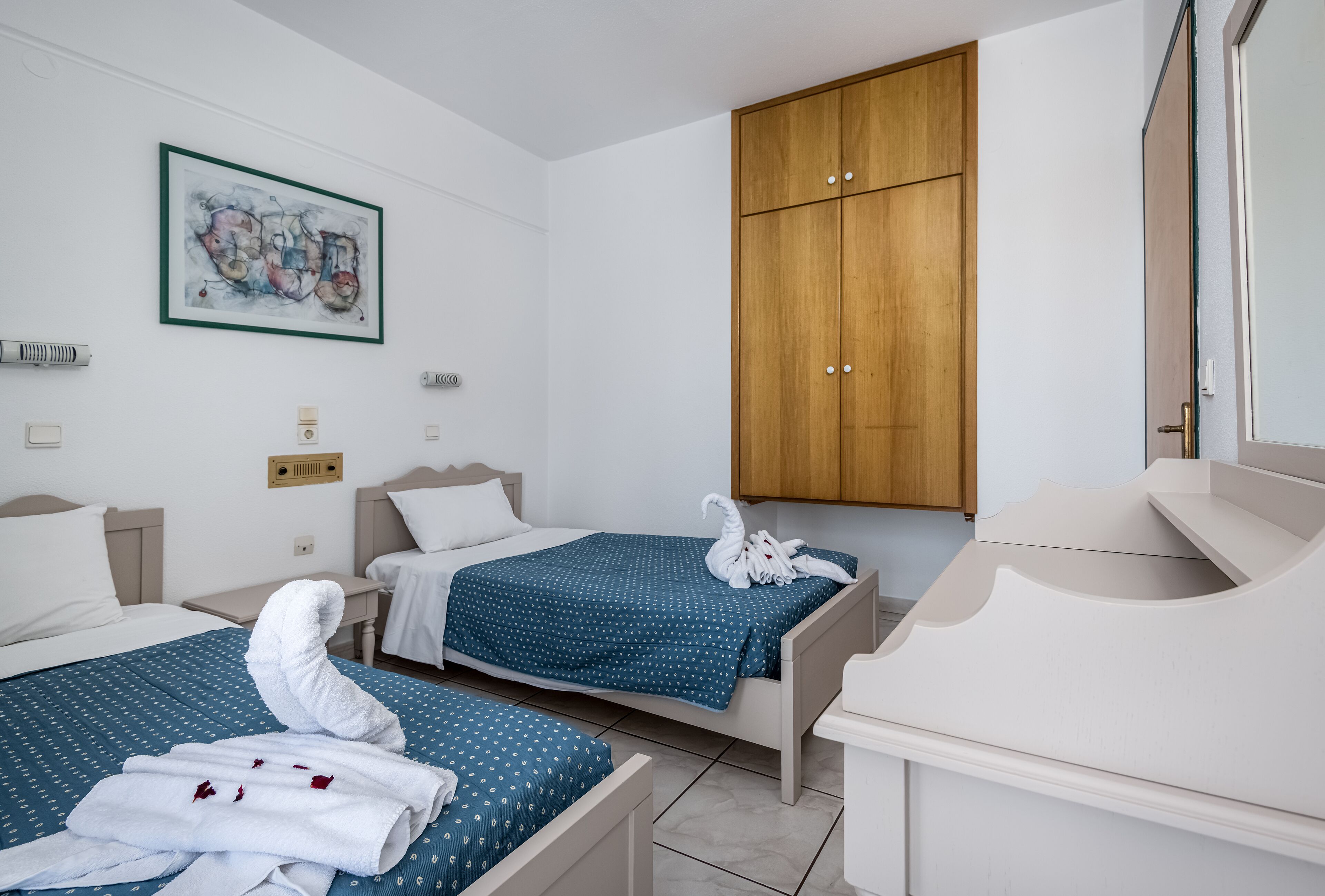Habitación de hotel con dos camas, un armario de madera, un cuadro en la pared y un baño con espejo y lavamanos.