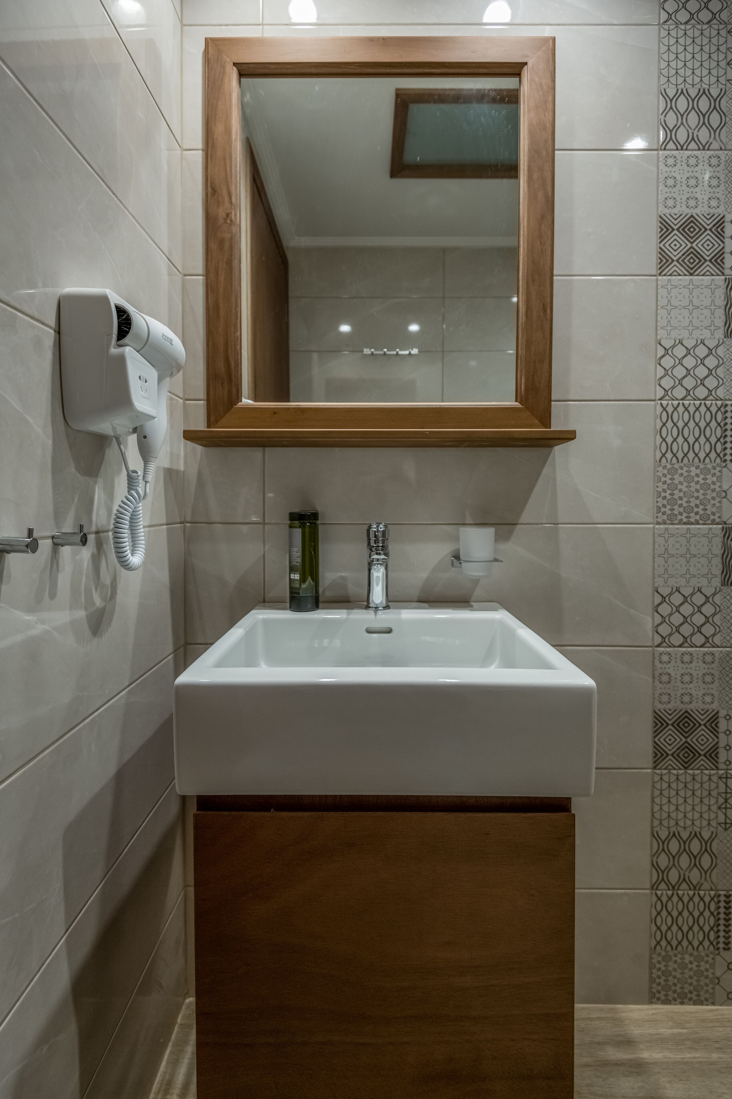 Un baño con lavamanos blanco, tocador de madera, espejo y secador de pelo en la pared.