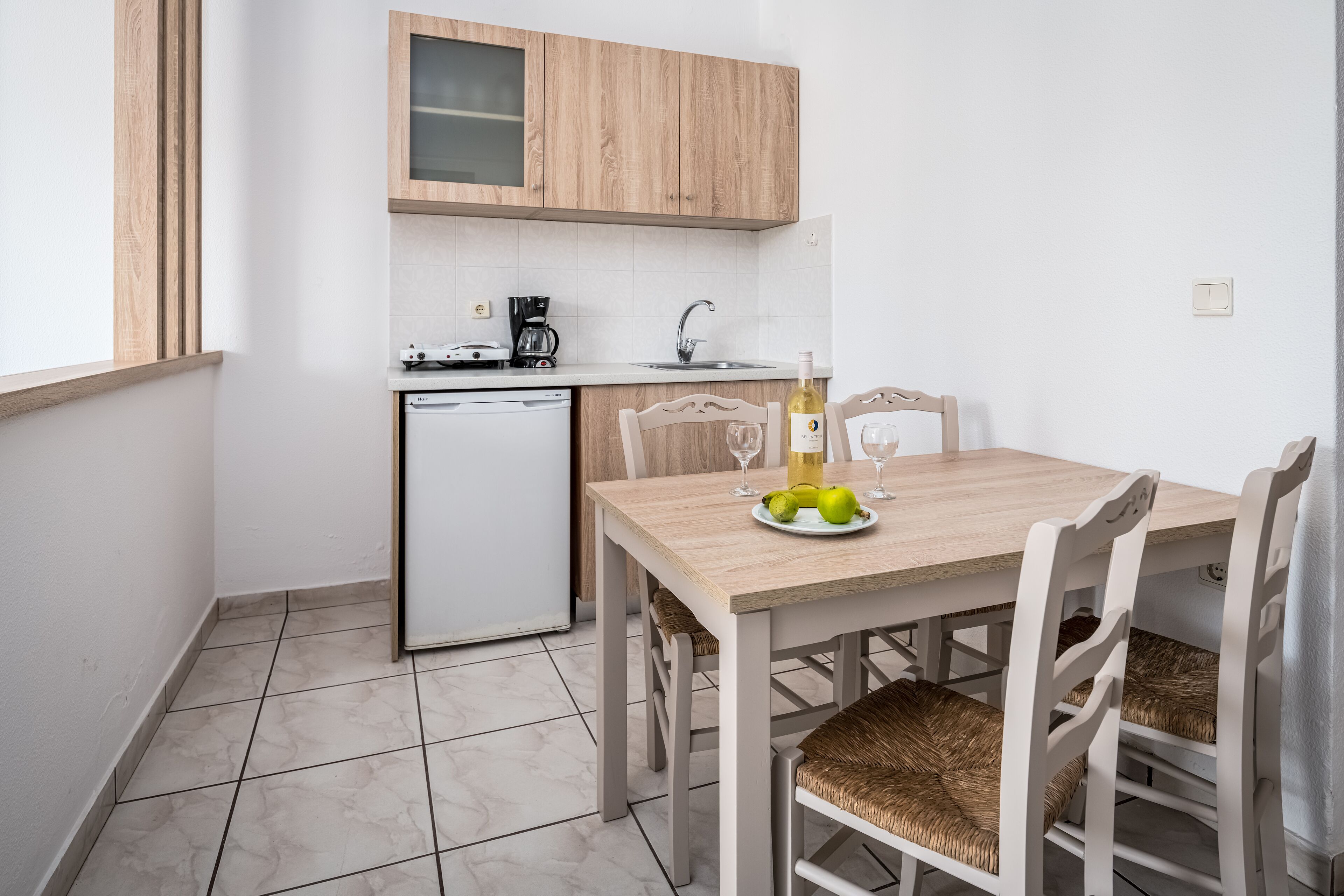 Cocina compacta con lavavajillas, cafetera y pileta. Hay una mesa de comedor con sillas y una botella de vino.