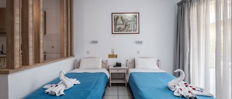 Habitación de hotel con dos camas, cada una con cubrecama azul y almohadas blancas. Hay un cuadro en la pared sobre las camas, una mesita entre ellas y una ventana con cortinas.