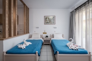A hotel room with two beds, each with a blue bedspread and white pillows. There is a painting on the wall above the beds, a small table between the beds, and a window with curtains.