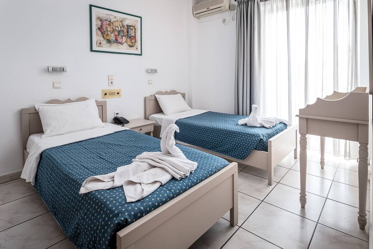 A hotel room with two beds, each with a blue bedspread and white pillows. There is a painting on the wall, a window with curtains, and a small table with a chair.