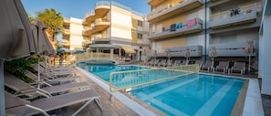 A swimming pool with sun loungers and umbrellas, surrounded by multi-story buildings with balconies.