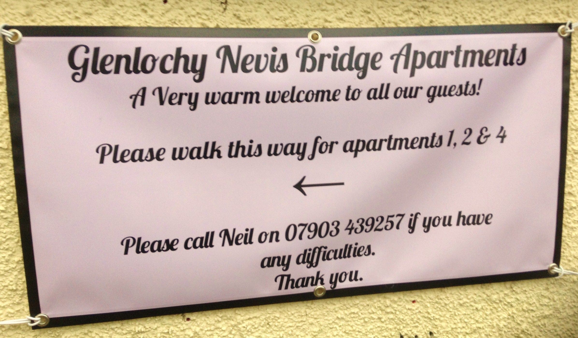 A welcome sign for Glenlochy Nevis Bridge Apartments, directing guests to walk a certain way for apartments 1, 2, and 4, and providing a contact number for assistance.