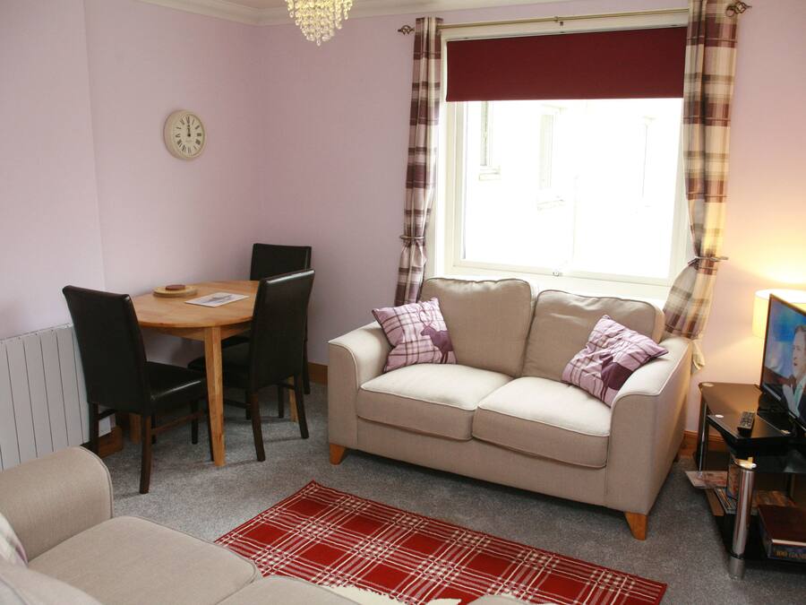 Un salon chaleureux avec un canapé beige, une table à manger avec des chaises, un tapis à motifs rouges et blancs, une horloge au mur et une fenêtre avec des rideaux.