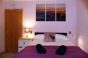 Een slaapkamer met een bed, een nachtkastje, een lamp, een deur en drie foto’s aan de muur.