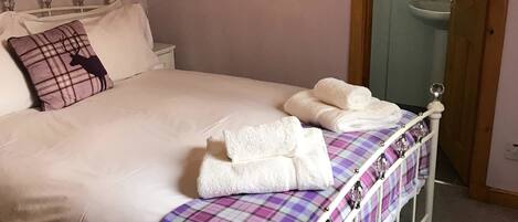 Un lit bien fait, avec du linge de lit blanc et une couverture à carreaux, une table de chevet avec une lampe, et une fenêtre donnant sur l’extérieur.