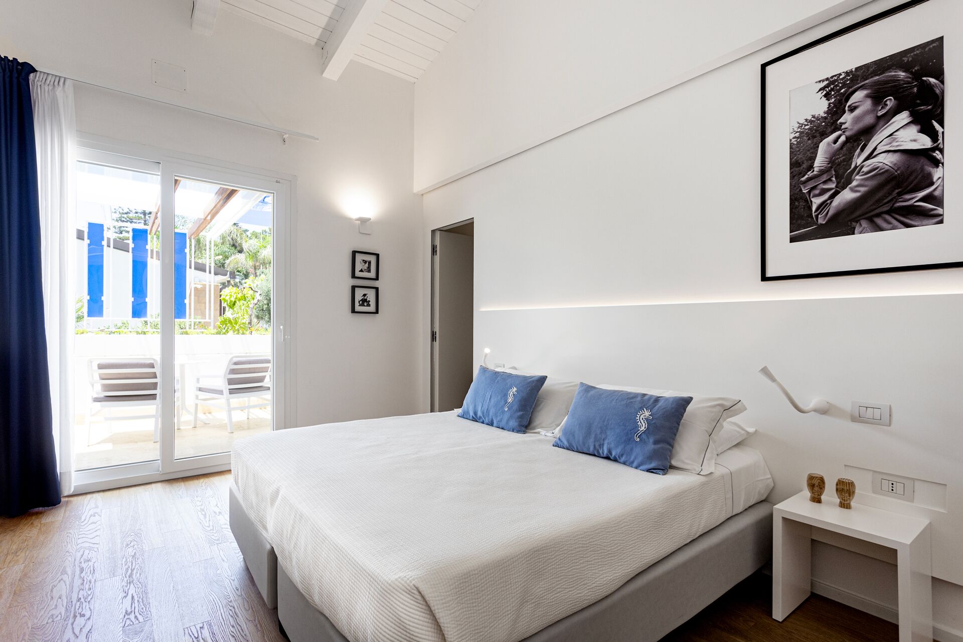 A modern bedroom with a large bed, a nightstand, a framed picture, and a view through a glass door.