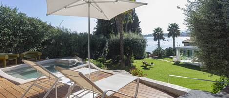 A wooden deck with lounge chairs and a large umbrella, overlooking a pool and a grassy area with a view of the sea.