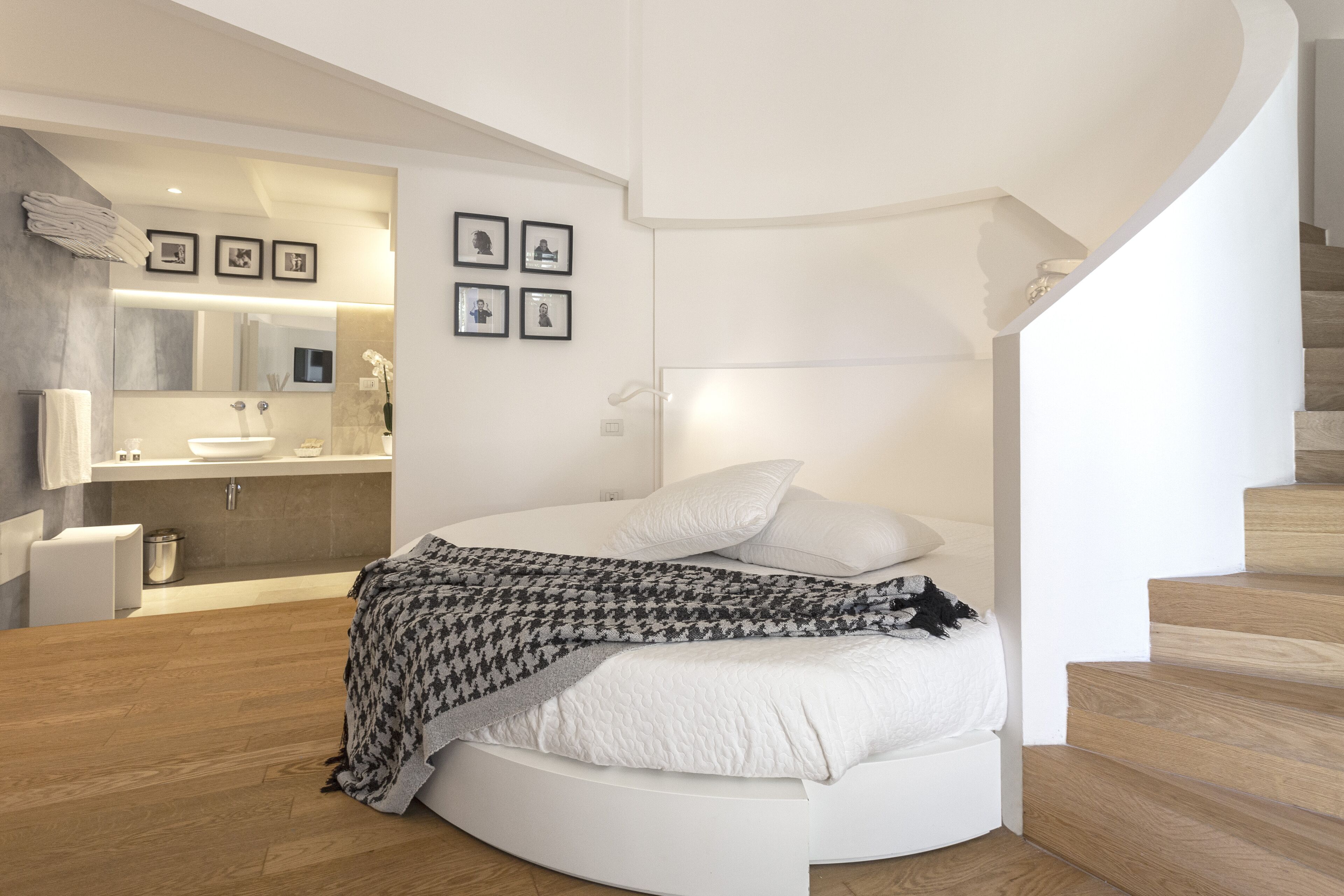 Quarto moderno com cama curva, banheiro integrado e escada.