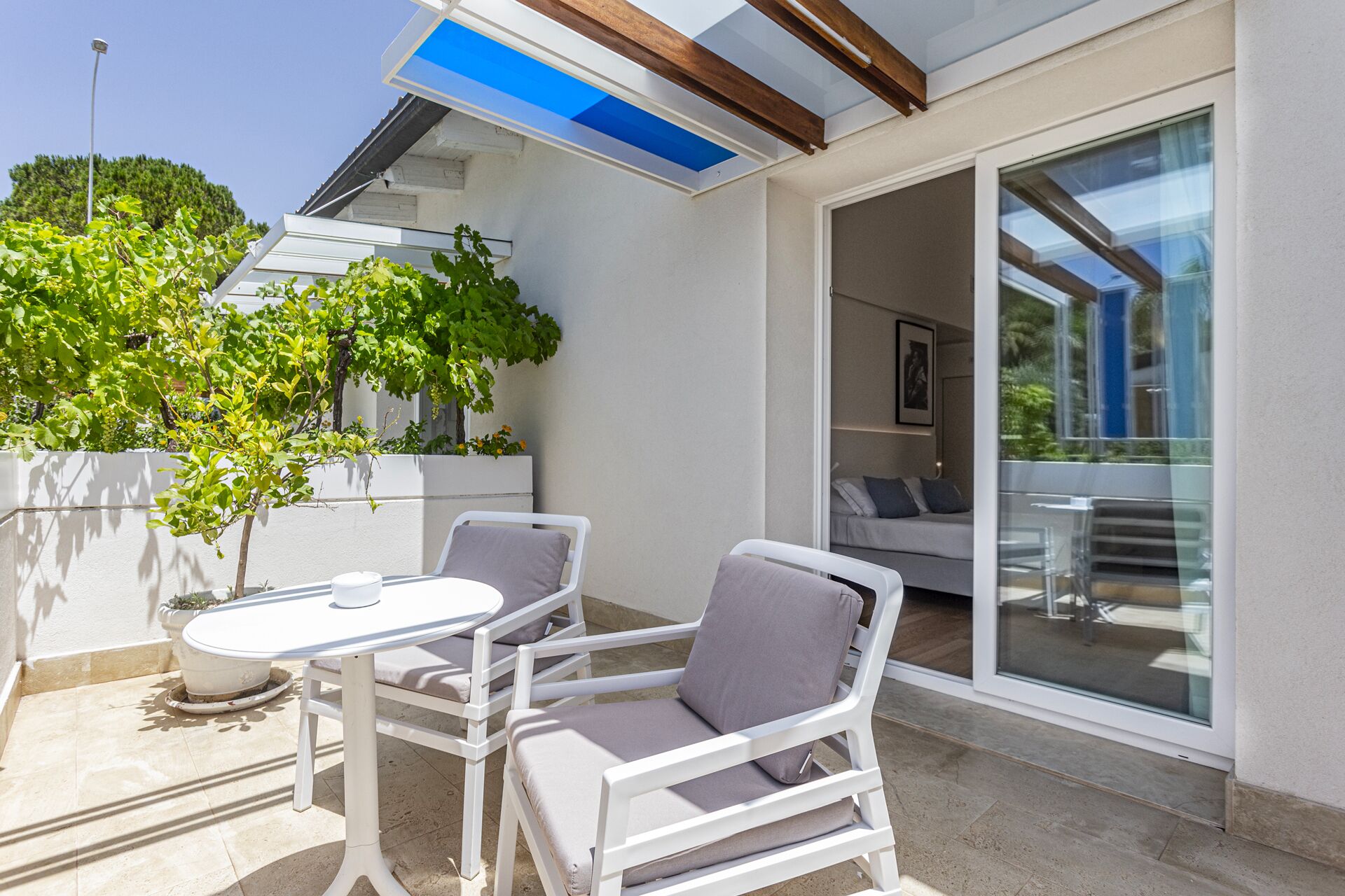 Eine Terrasse mit zwei weißen Stühlen und einem Tisch, eine Glastür, die zu einem Innenbereich führt, und ein kleiner Baum mit grünen Blättern.