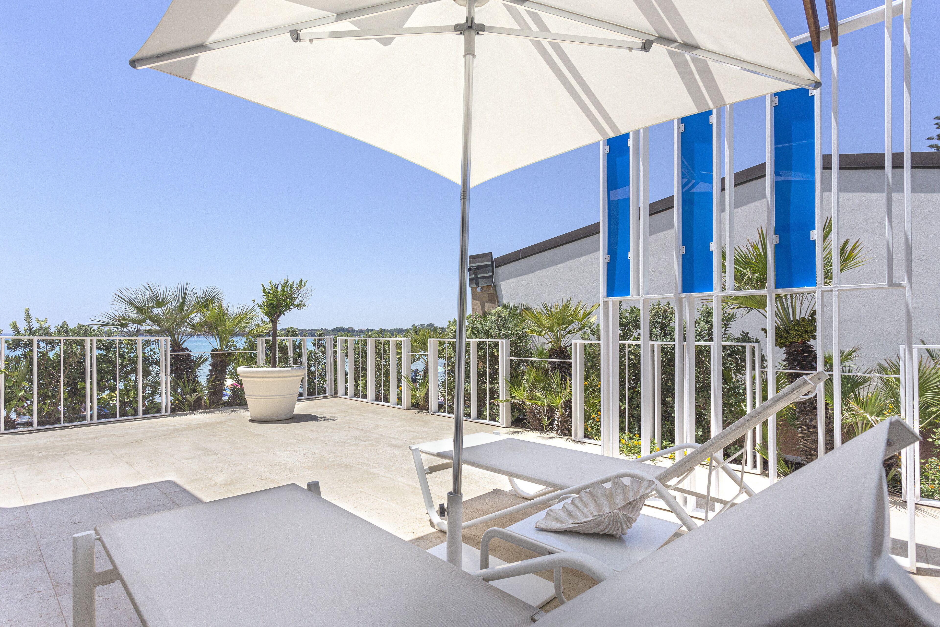 A sunlit outdoor seating area with white lounge chairs and a large umbrella, overlooking a body of water and a modern building with blue accents.