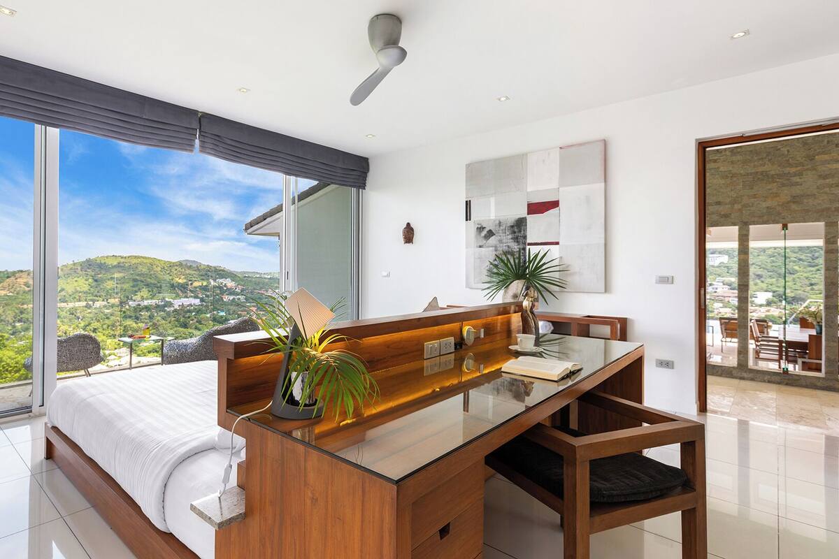 A modern bedroom with a large bed, a wooden desk, and a view of a green landscape.
