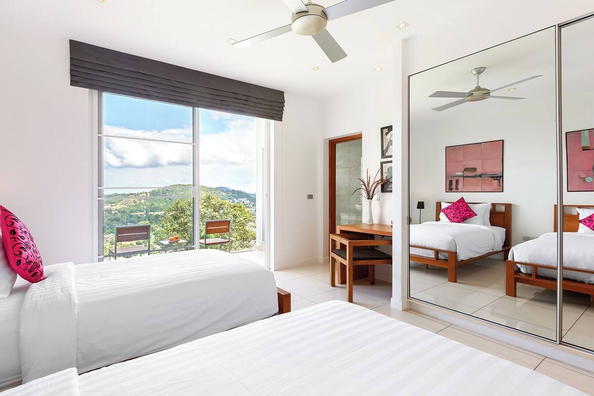 A hotel room with a large bed, a desk, and a chair. There is a window with a view of the outdoors.