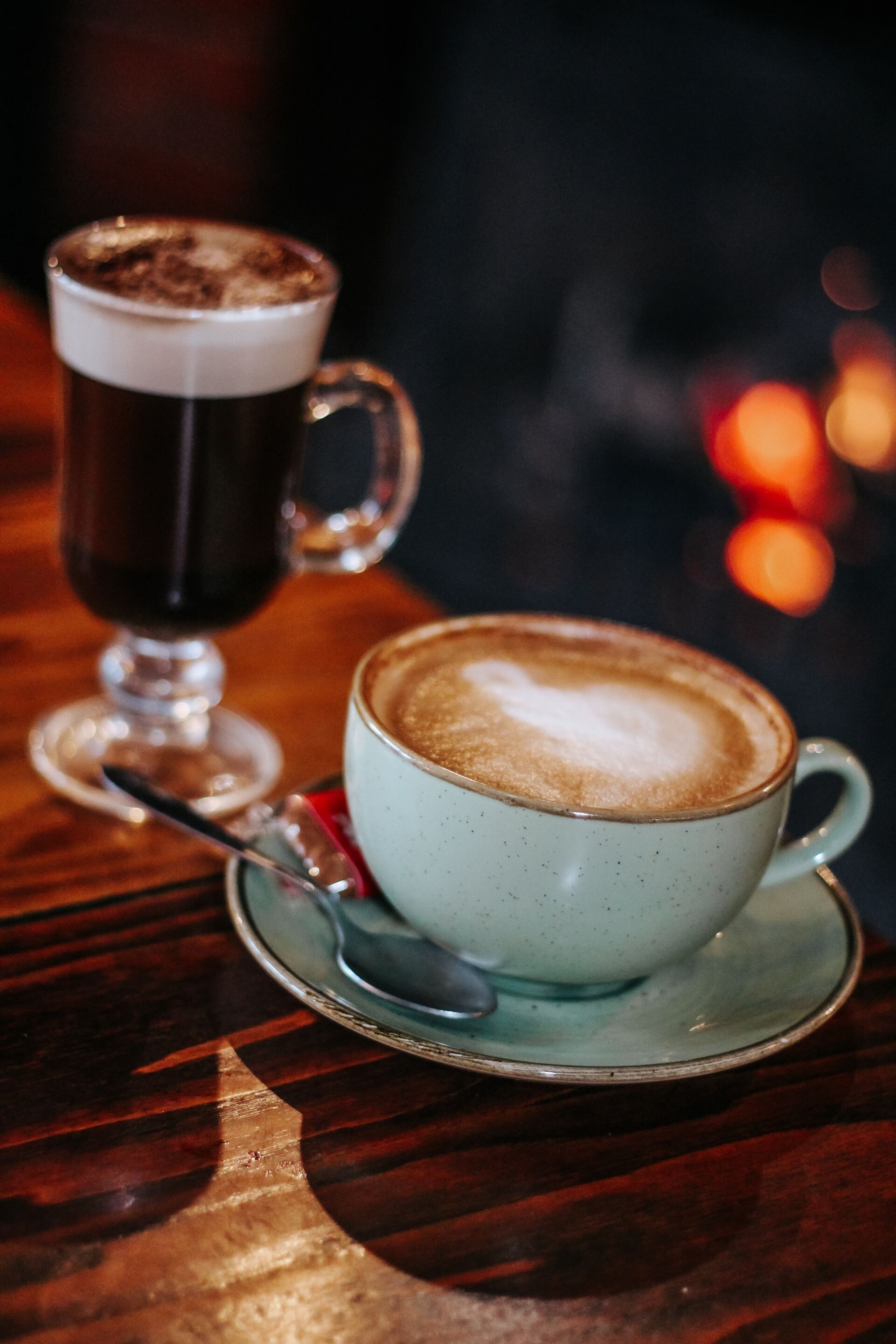 A cup of coffee with a frothy top on a saucer, a spoon, and a glass with a similar beverage.