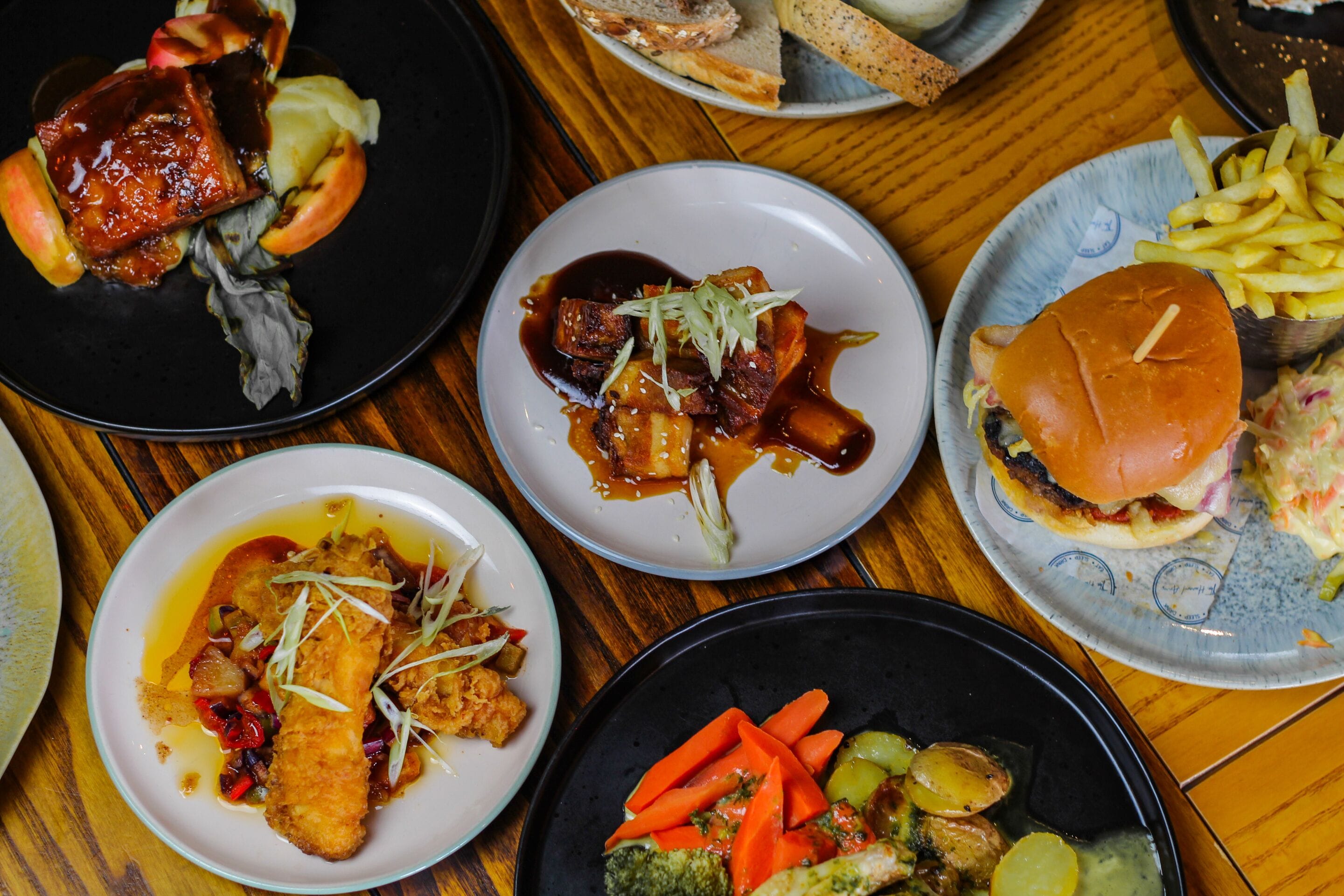 On peut voir une variété de plats sur une table en bois, notamment un hamburger, des frites et une assiette contenant ce qui semble être un plat de poisson.