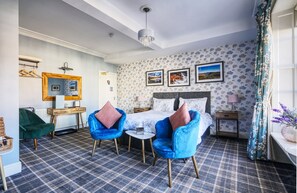 A hotel room with a checkered floor, a bed with floral wallpaper, a desk with a mirror, and two blue chairs. - Howard Arms (Brampton)