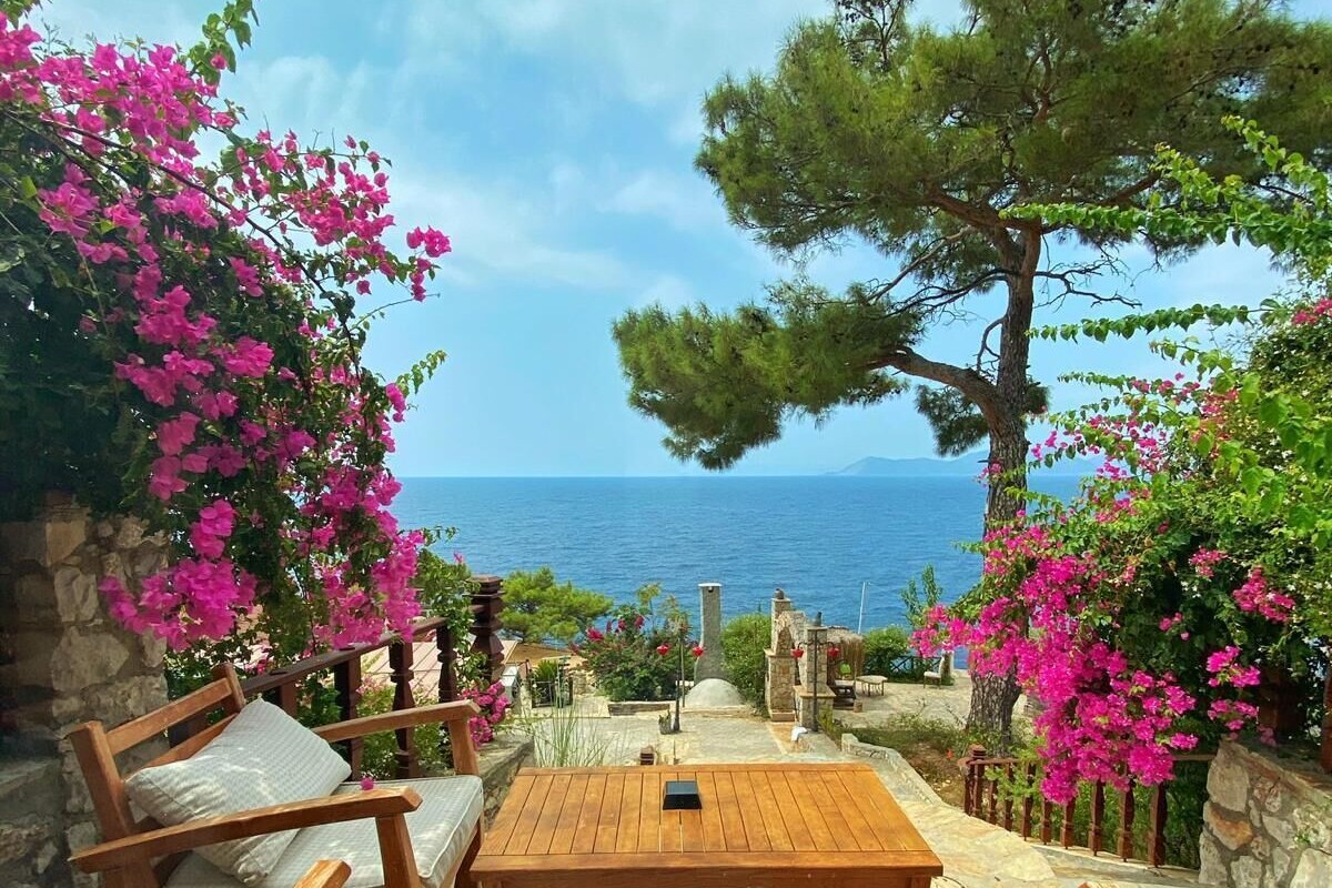Zona salotto esterna in legno con parete in pietra, vista sul mare e piante fiorite.