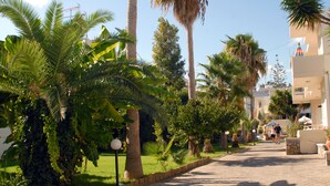 A paved pathway lined with palm trees and greenery, leading to a building with balconies and outdoor seating. - Evilion Hotel (Chania)