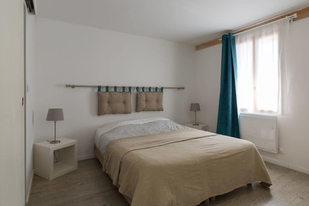 Une chambre à coucher avec un lit, des tables de chevet, une fenêtre avec des rideaux et une tête de lit agrémentée d’oreillers.