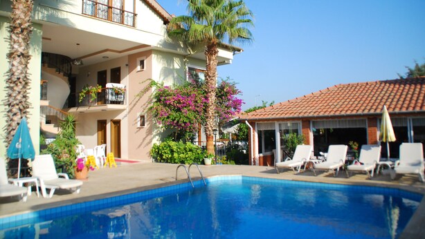 Zona junto a la piscina con una piscina azul cristalina, sillas de descanso blancas y un edificio de dos pisos con balcones.