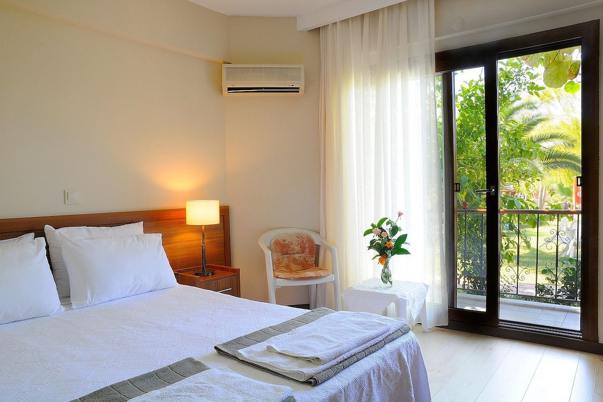 A hotel room with a bed, a chair, a table with a vase, and a balcony with a view of greenery.