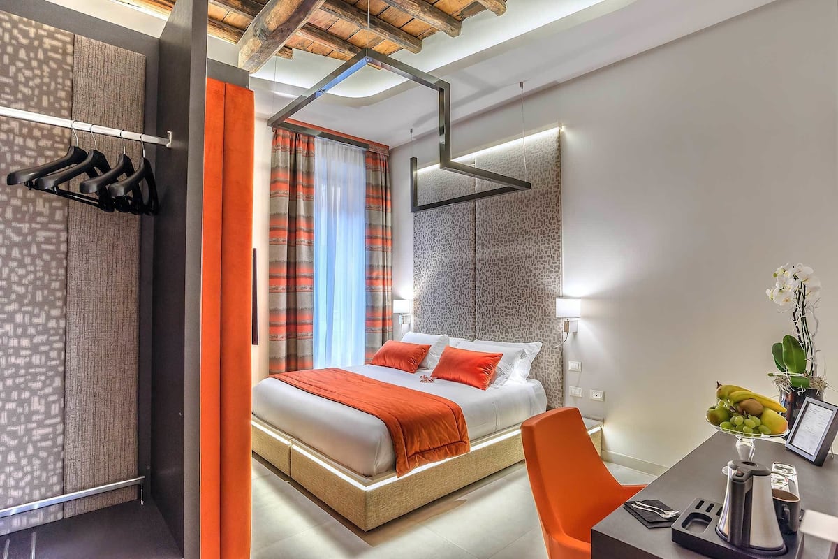 A modern hotel room with a large bed, orange curtains, a desk with a coffee maker, and a view of the city.