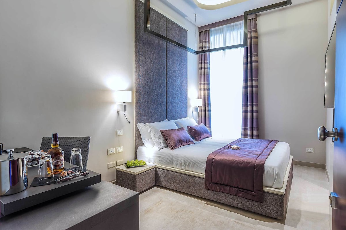 A modern hotel room with a large bed, a bedside table with a bottle and glasses, and a small side table with fruits.