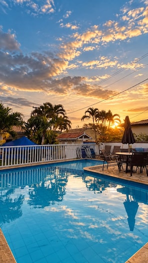 A swimming pool with lounge chairs and umbrellas, surrounded by palm trees and a house with a tiled roof. - Pousada Martito Geriba (Búzios)