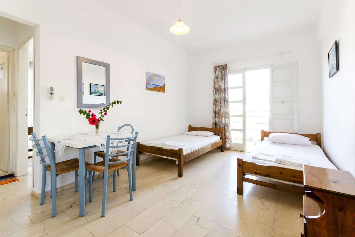 A small, clean room with a wooden bed, a small table with chairs, a mirror, and a window with curtains.