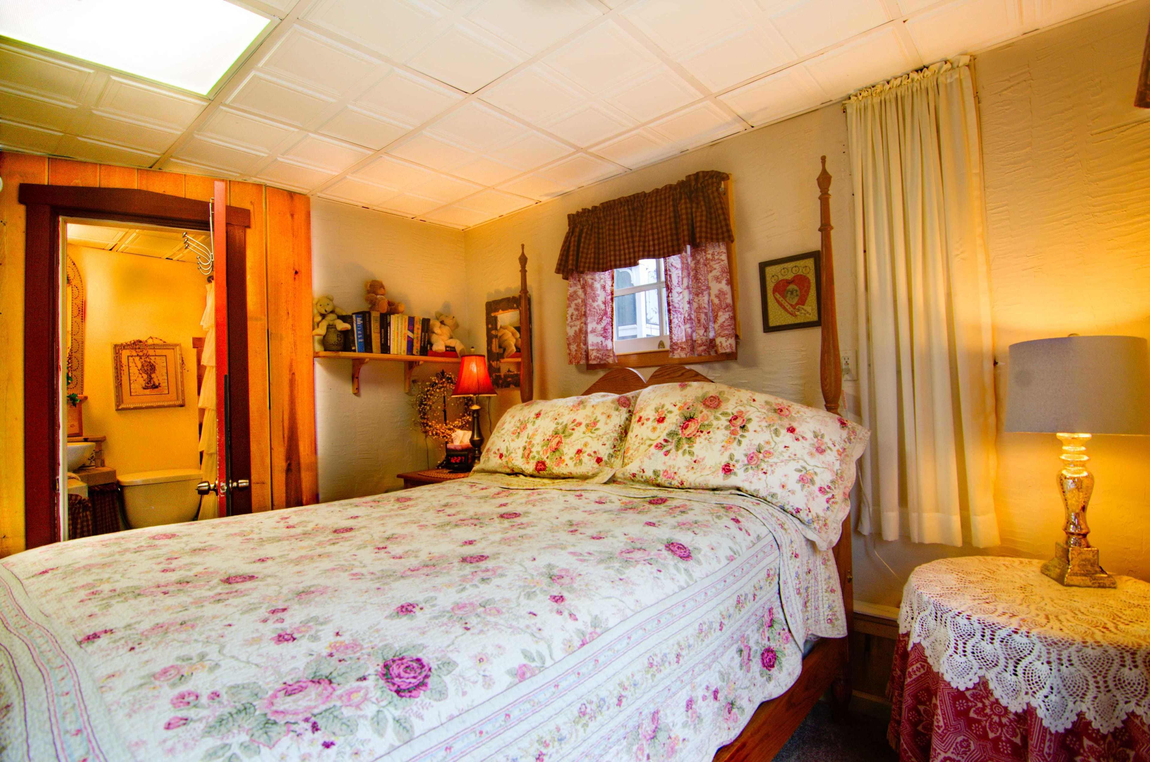 A bedroom with a floral bedspread, a bedside table with a lamp, a window with curtains, and a wooden door leading to another room.
