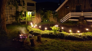 Een rustieke schuur met een witte omheining, een houten trap en een goed verlichte tuin met lichtjes.