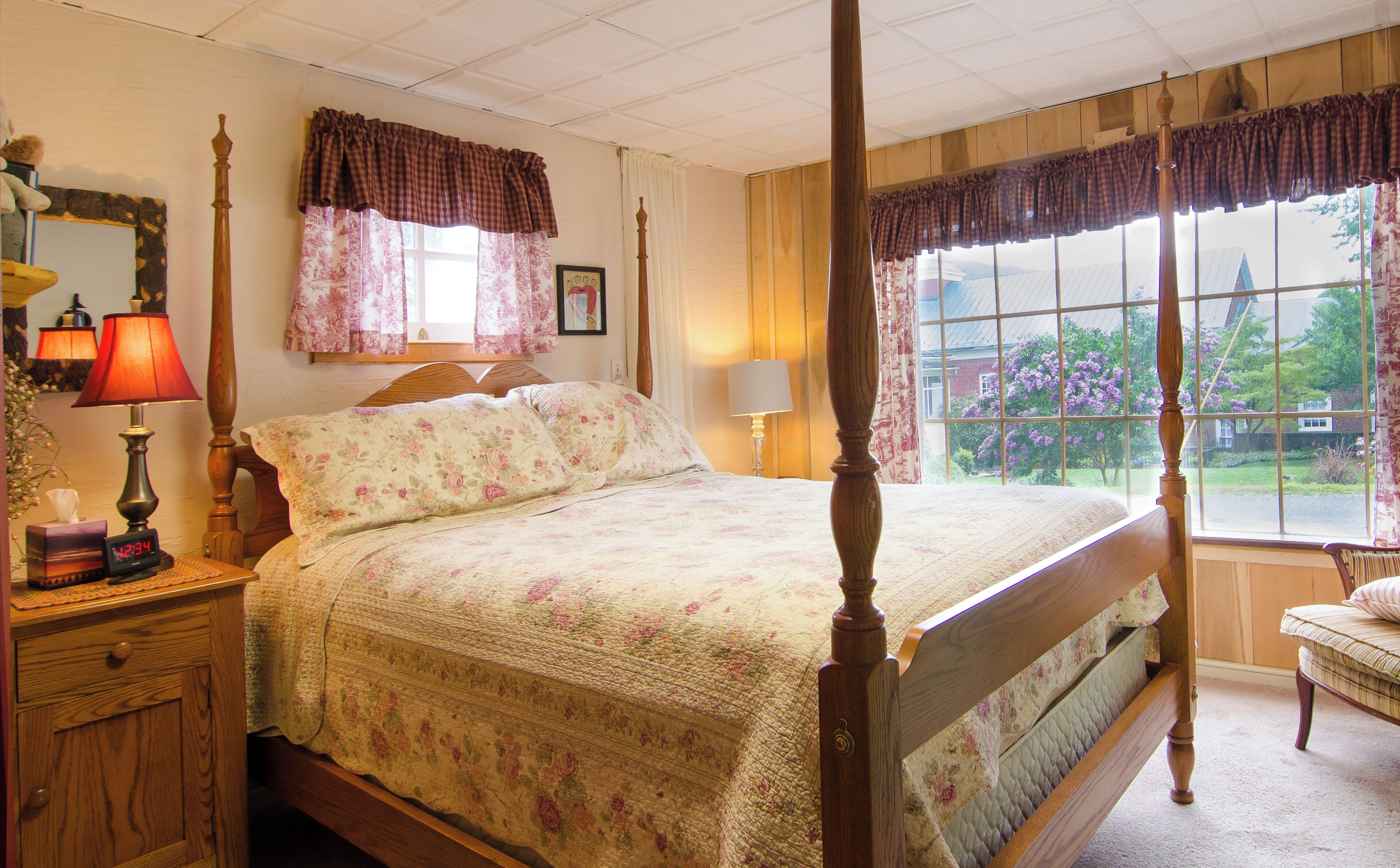 Una cama con dosel, ropa de cama floral, una mesita de noche con lámpara y vistas a un jardín a través de un ventanal grande.