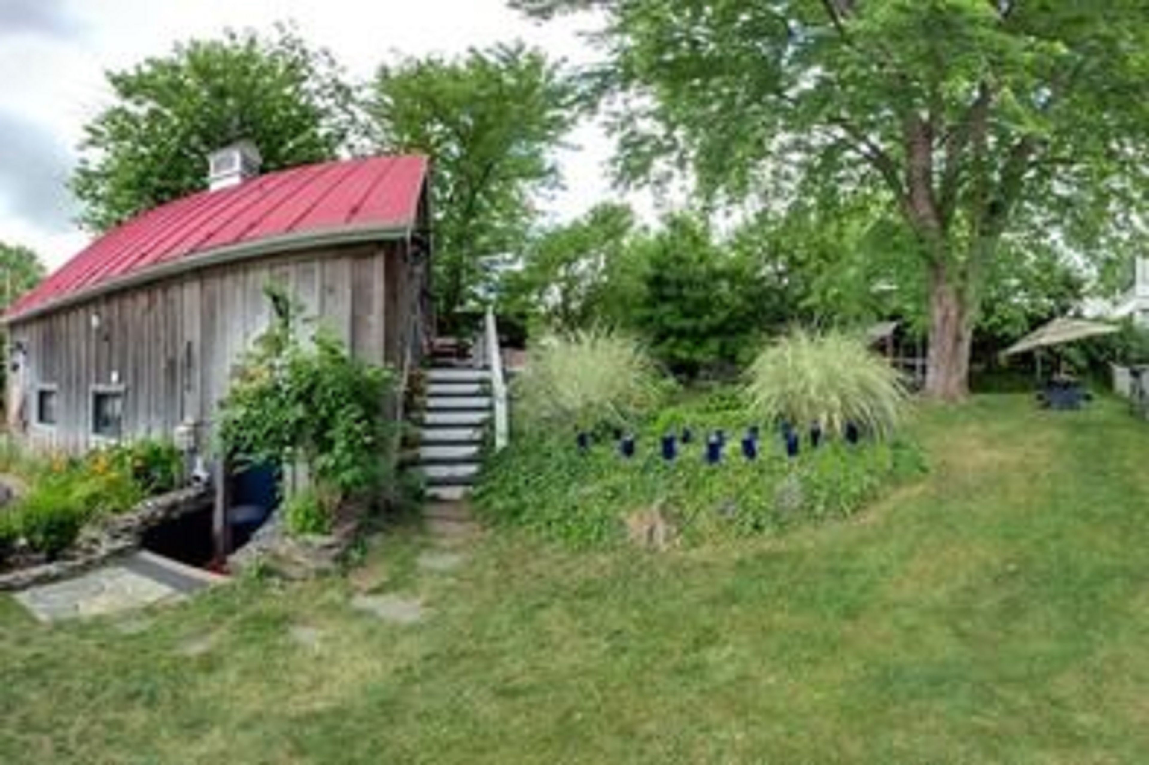 Une grange rustique en bois, avec un toit rouge, entourée d’une végétation luxuriante et accessible par un escalier en pierre.