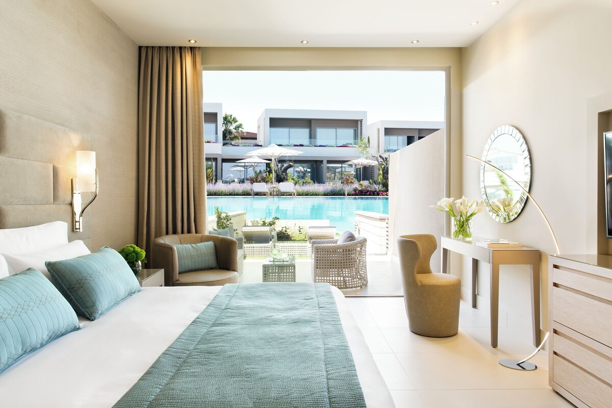 A modern hotel room with a large bed, a view of a pool, and a balcony with outdoor furniture.
