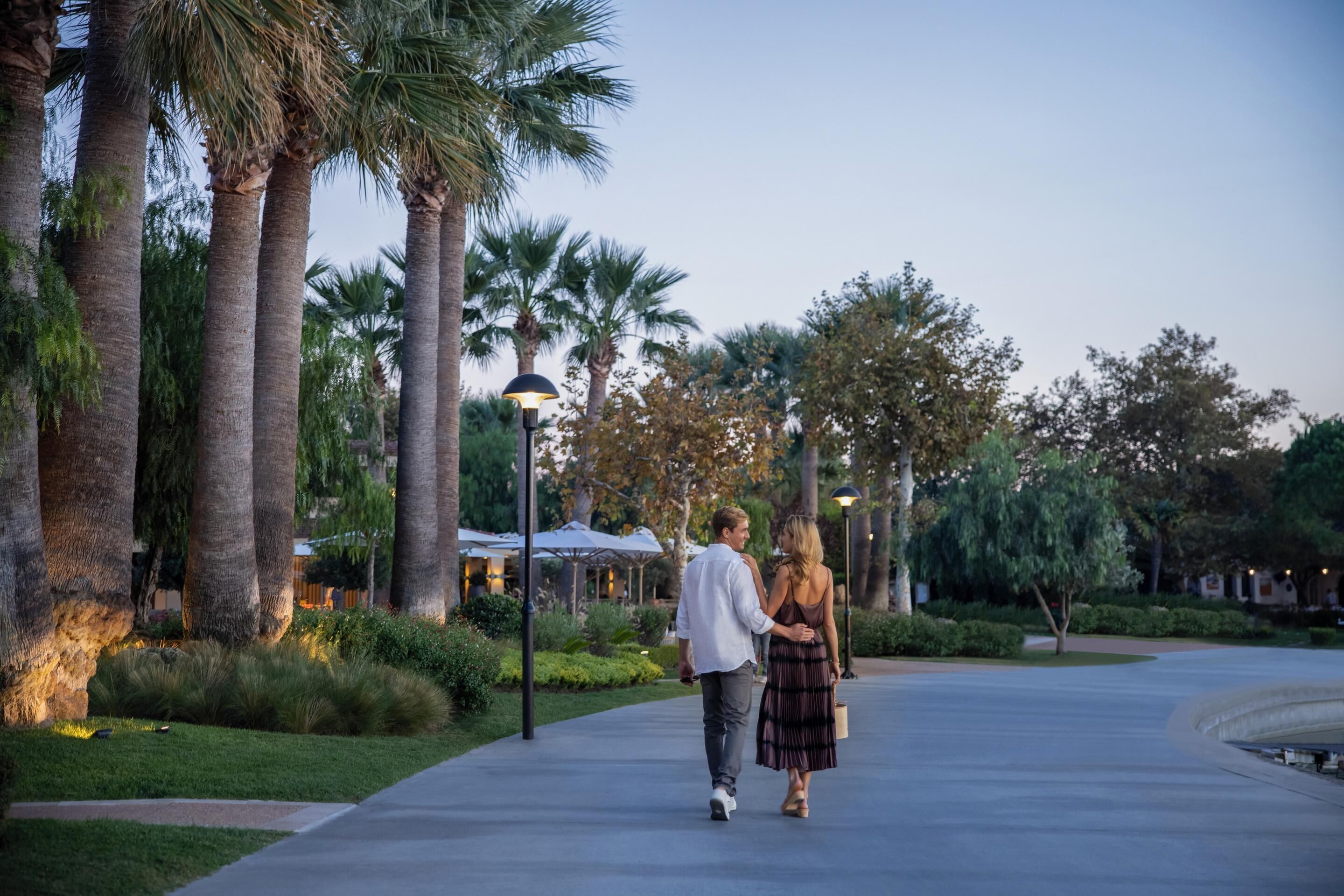 A couple walking on a paved path surrounded by palm trees and greenery, with outdoor lighting and a building in the background.