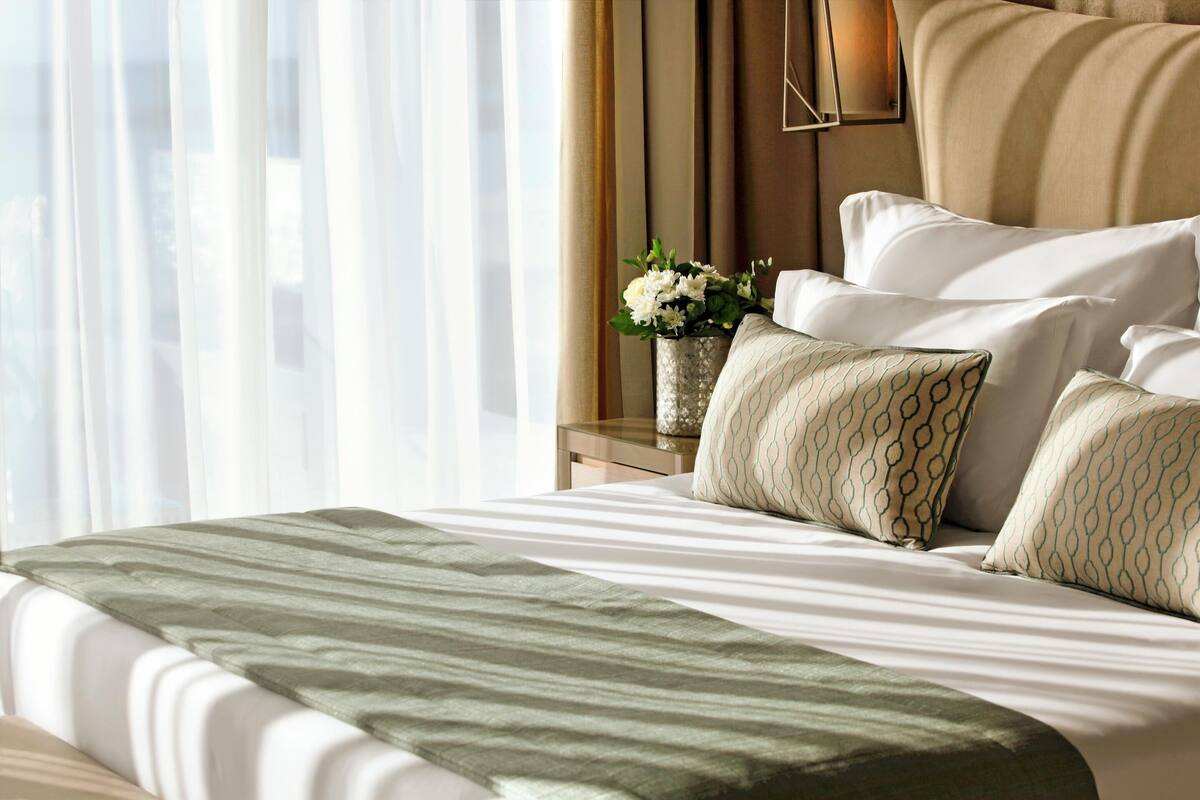A neatly made bed with patterned pillows, a green throw blanket, and a vase of flowers on a bedside table.